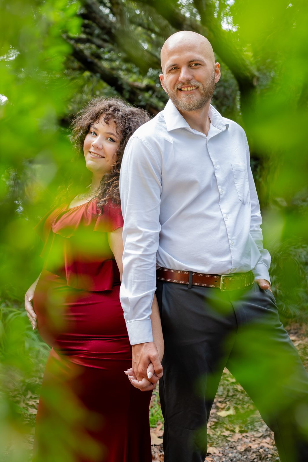 Maternity couple in red gown surrounded by greenery