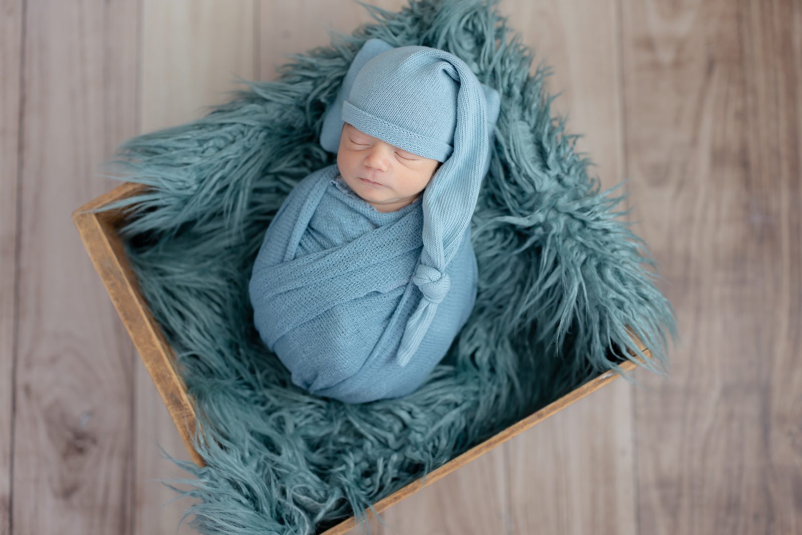 Newborn swaddled in dusty blue with a sleepy cap, nestled in a wooden crate on blue fur