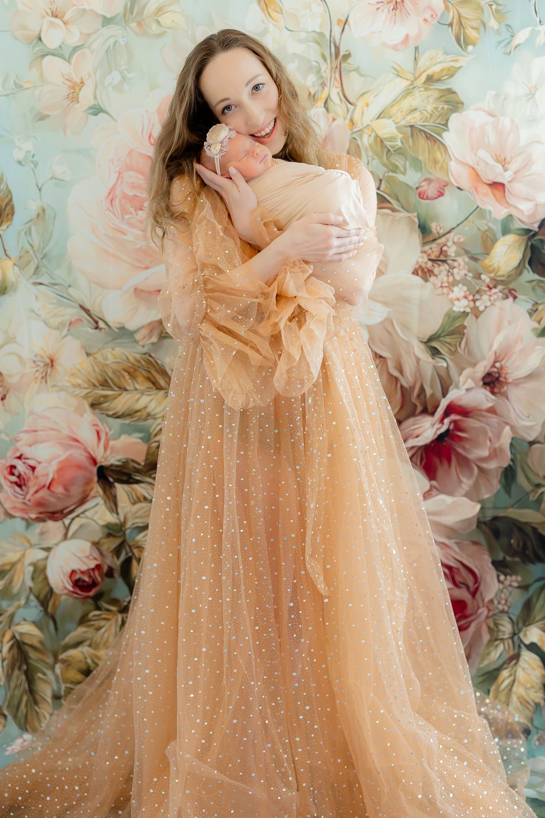 Mother in a peach gown holding her newborn against a floral backdrop