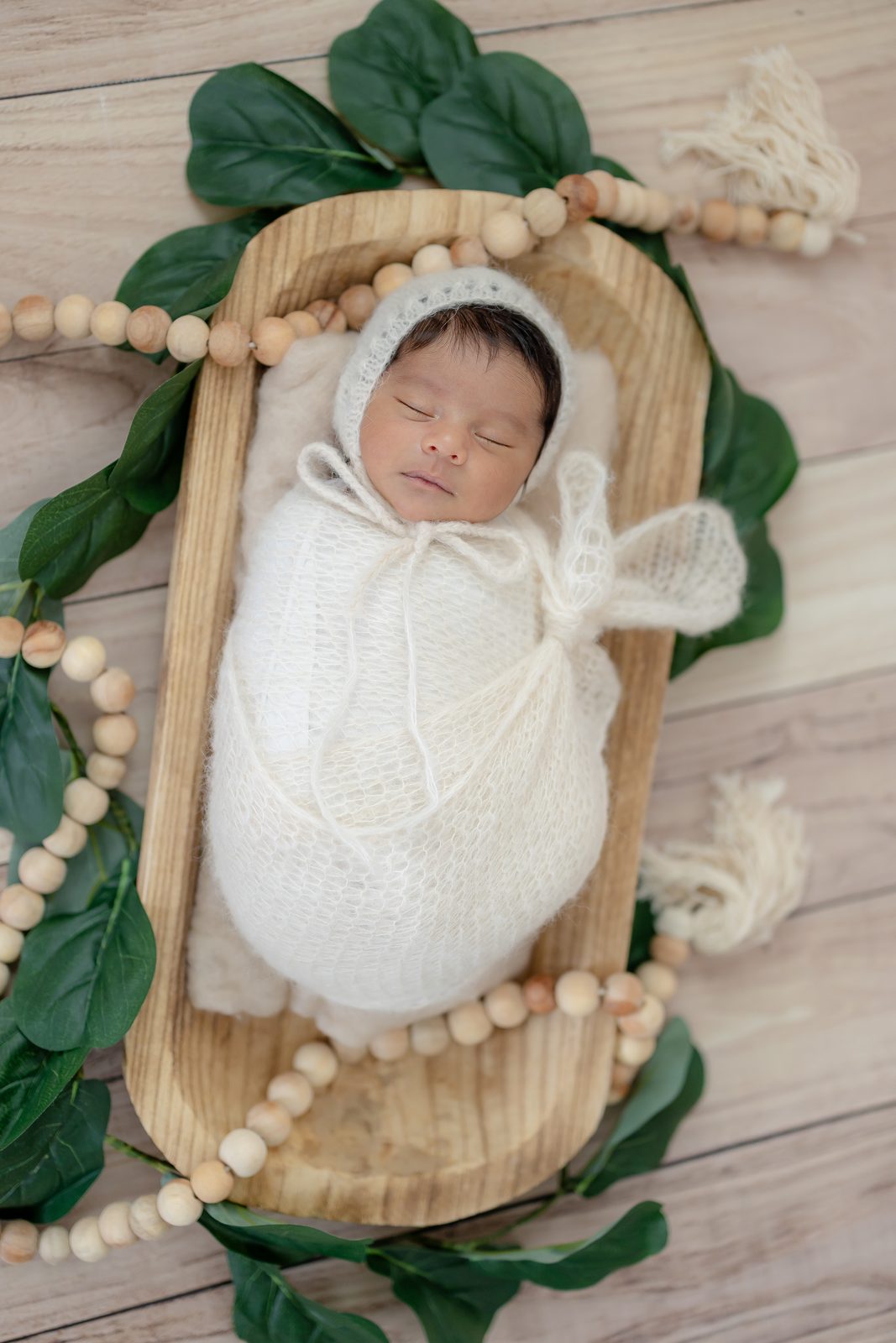 Newborn in a white bonnet resting in a wooden bowl with greenery and wood beads