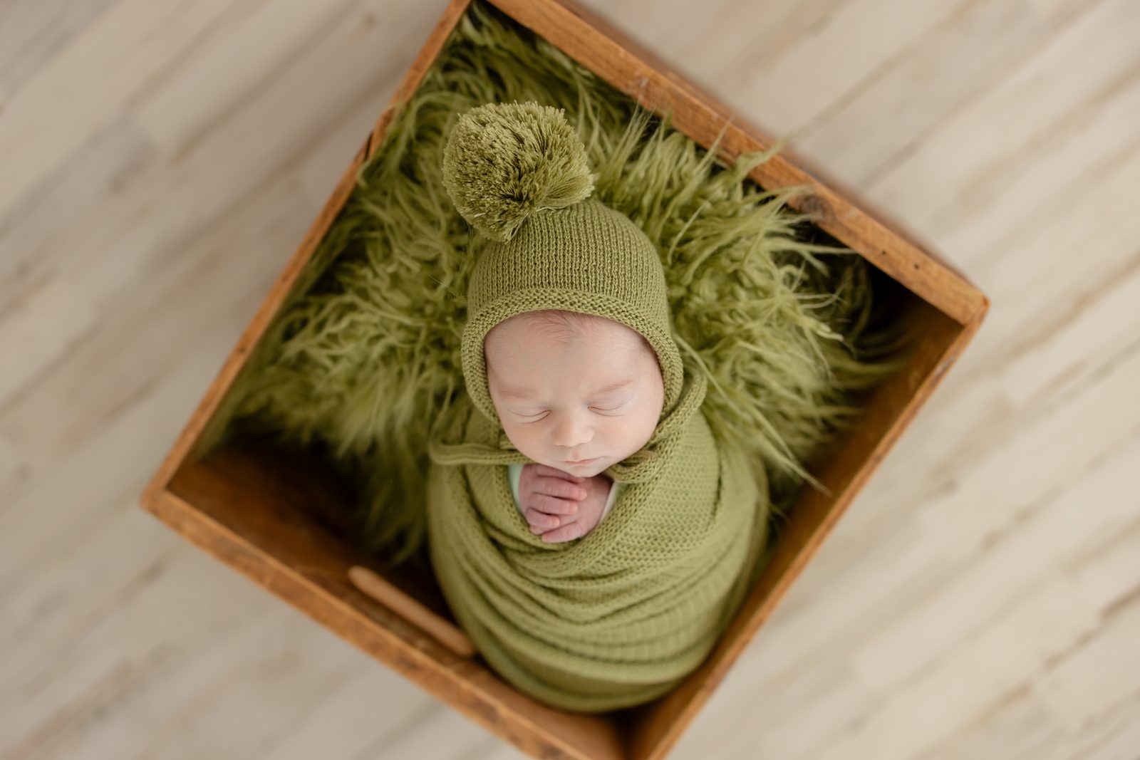 Newborn swaddled in olive green with a pom-pom bonnet, nestled in a wooden crate
